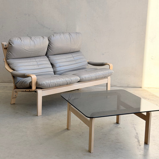 The Tessa Ferrara Lounge Suite – Complete 5-Piece Set (Revive), featuring modern light wood frames and gray cushions, sits against a plain wall next to a rectangular glass coffee table with matching wooden legs on a light floor.