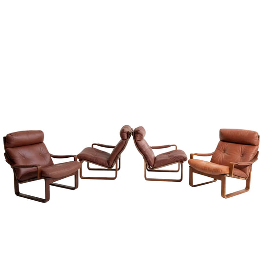 Four Tessa T8 Lounge Suite modular two-seater sofas and armchairs (Revive) in brown leather with wooden frames, inspired by mid-century modern design, are arranged in a minimalist, sunlit room with light grey walls and a pale floor. A wooden cabinet sits in back.