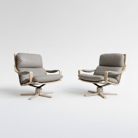 A pair of Tessa Ferrara Swivel Armchairs (Revive), featuring light wooden frames and grey cushioned seats, are positioned side by side against a plain light wall on a concrete floor.