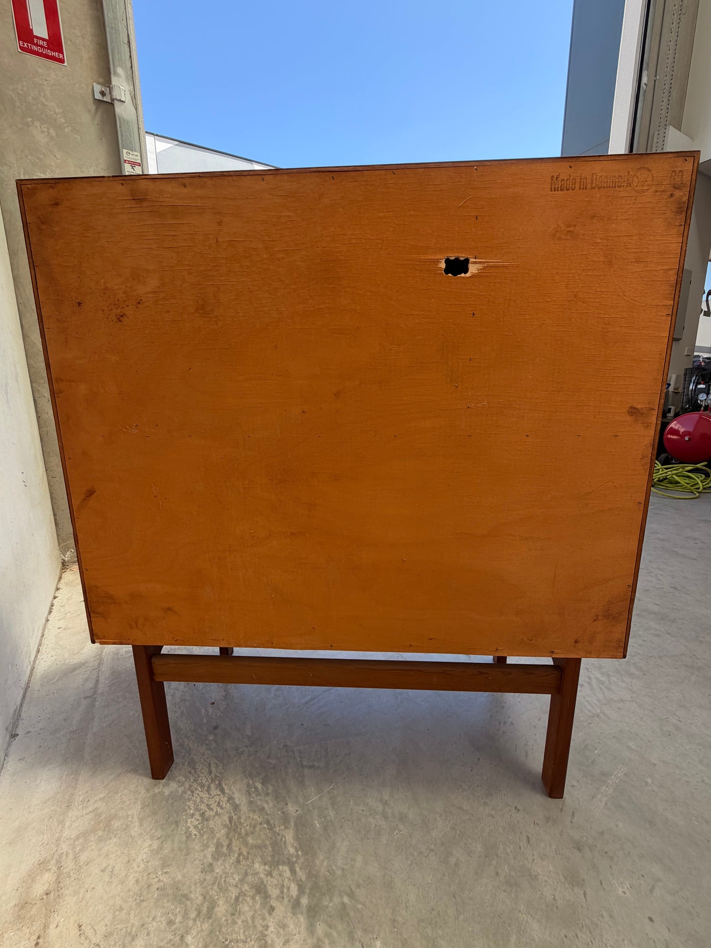 A Mid Century Modern Danish Arne Wahl Iversen Model 68 Teak Secretary Desk by Vinde Møbelfabrik stands on a concrete floor, its back visible with a small hole and "Made in Denmark" stamped above; industrial equipment appears in the background.