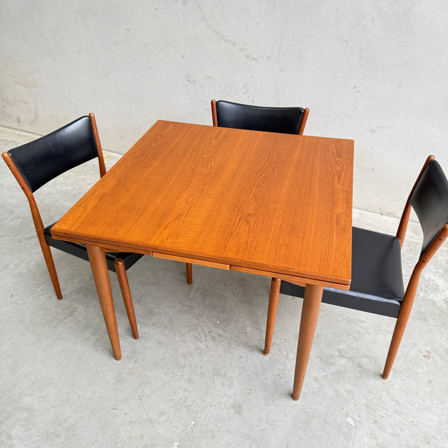 The Fully Restored Parker 140 Square Extension Table (Rediscover), featuring a mid-century modern design with tapered legs, is paired with three black wooden dining chairs on a light gray concrete floor against a plain gray wall.