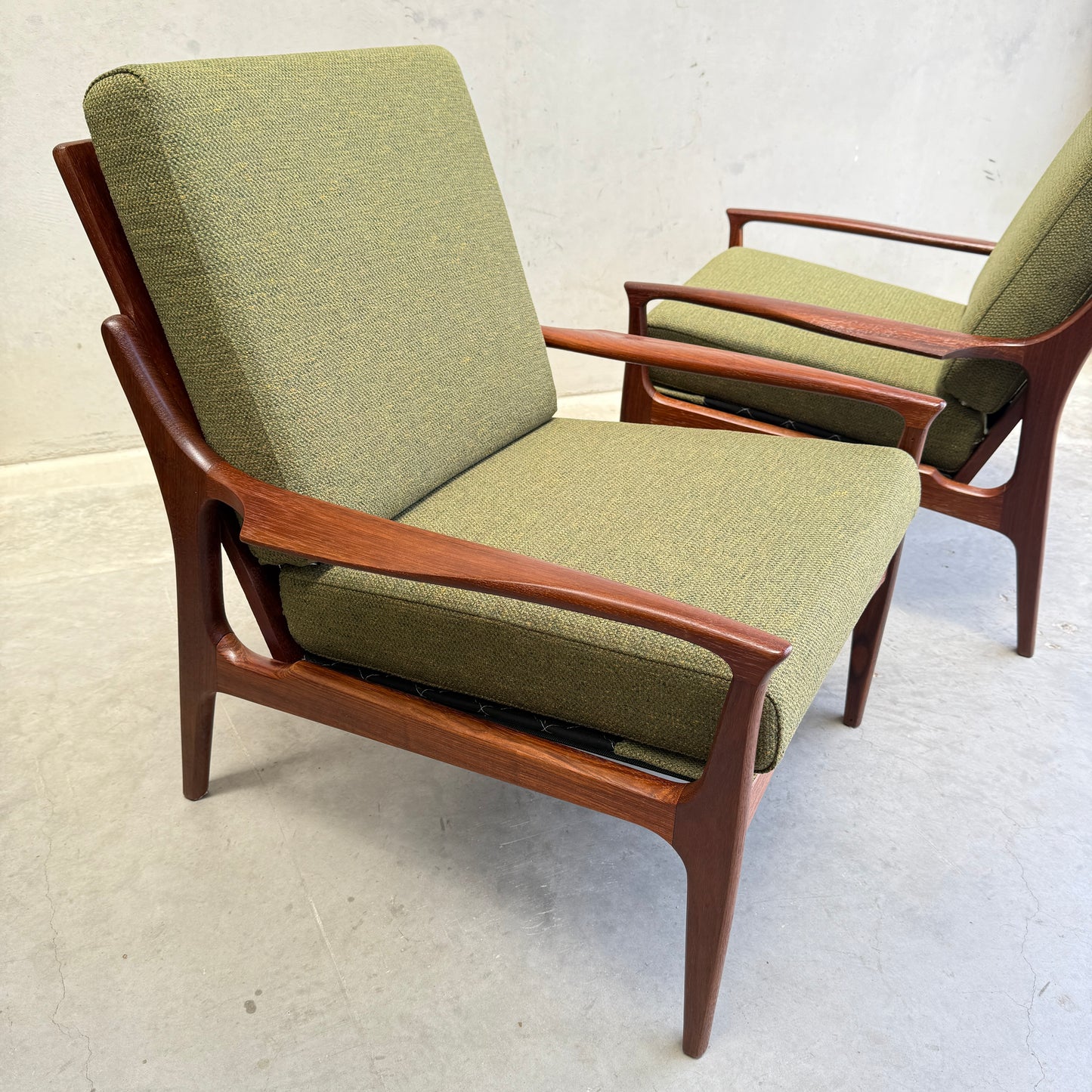 Two Don Rex Armchair - Green Forest (Rediscover) with upholstered green cushions and wooden frames sit on a light concrete floor against a plain wall, showcasing the elegance of mid-century Australian design.