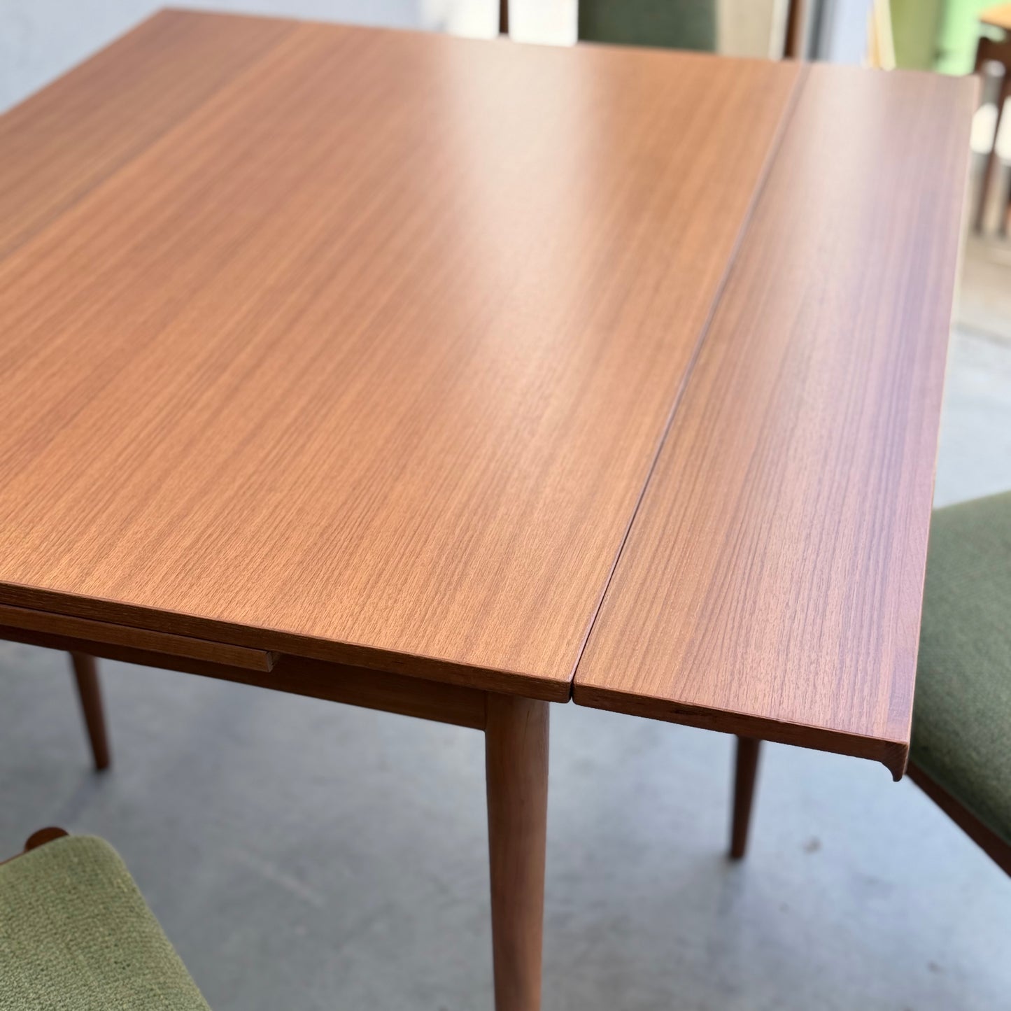 A close-up of the Fully Restored Parker Dining Suite 5 Piece - Forest Green (Rediscover), featuring a Scandinavian-style wooden table with an extended leaf and green cushioned chairs on a smooth, light floor.