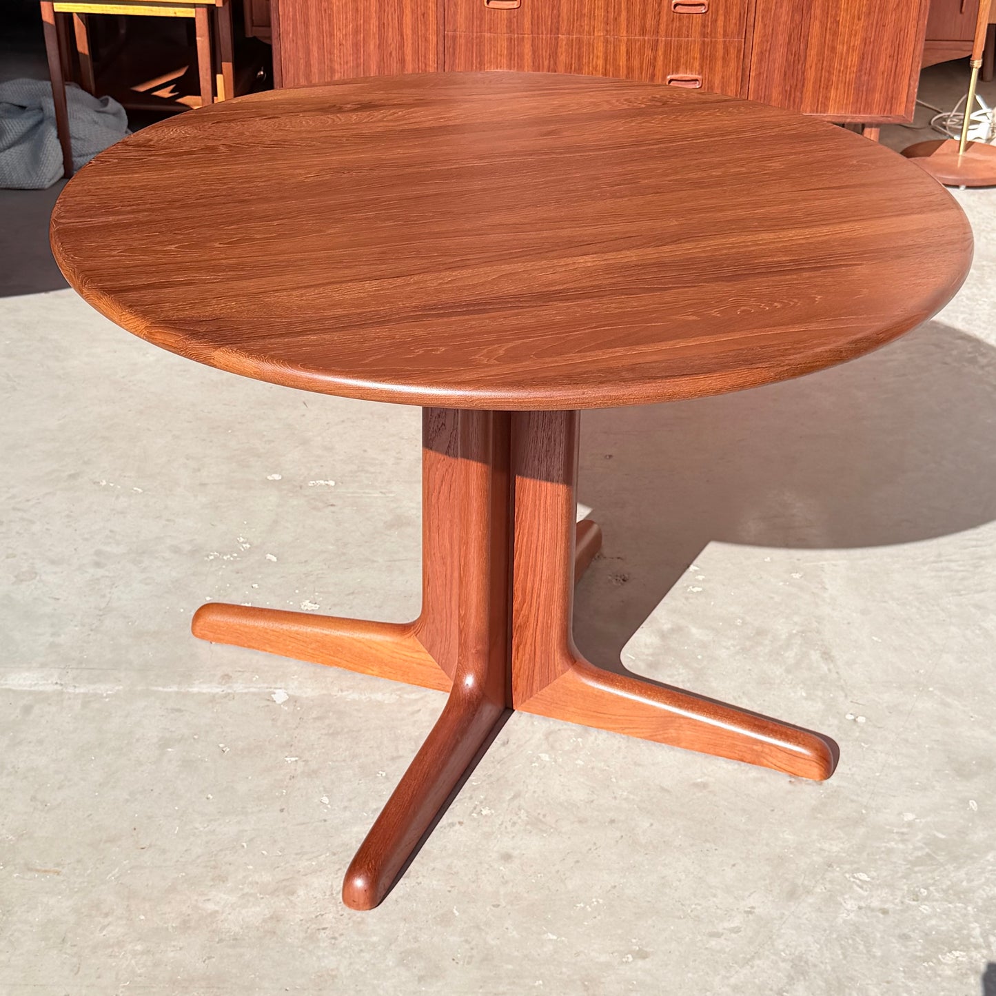 The Fully Restored Solid Teak Danish Dining Table (Rediscover) features a vintage round design with a smooth finish and central pedestal base splitting into three curved legs, set on a light-colored concrete floor.
