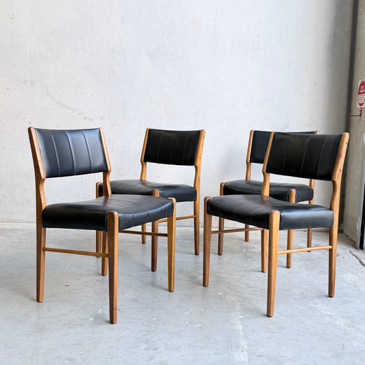 A set of four Vintage TH Brown Blackwood Dining Chairs (Revive) with black cushioned seats and backs, showcasing a refined mid-century modern design, are arranged in a spacious room with light gray concrete walls and floor.