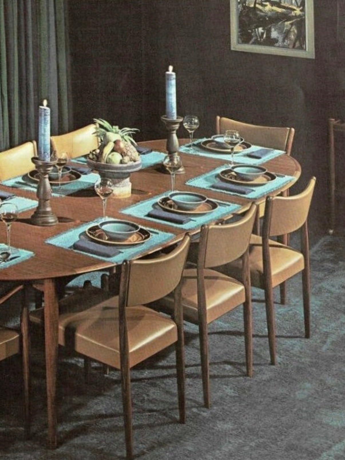 A mid-century modern dining room features a teak table set for six with Parker Number 97 Dining Chairs Set of 4 – Licorice Vinyl, blue placemats, wine glasses, tall blue candles, and a fruit centerpiece. Dark walls and carpeted floor add cozy elegance.