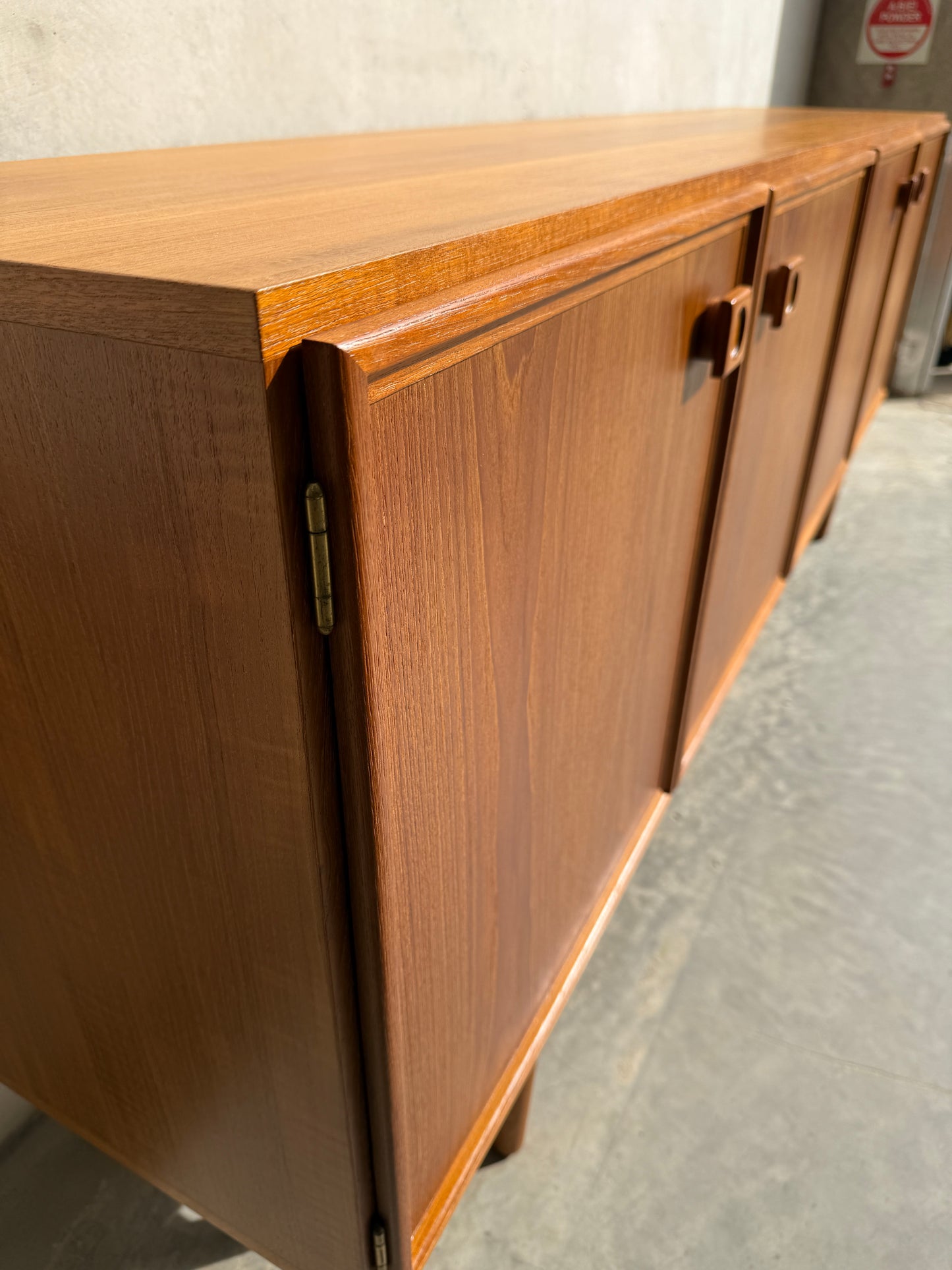 A close-up of the Iconic Parker Teak 4-Door Sideboard – Restored Mid Century Modern with tapered legs, highlighting its panel doors and geometric handles, set on a concrete floor against a plain light wall.
