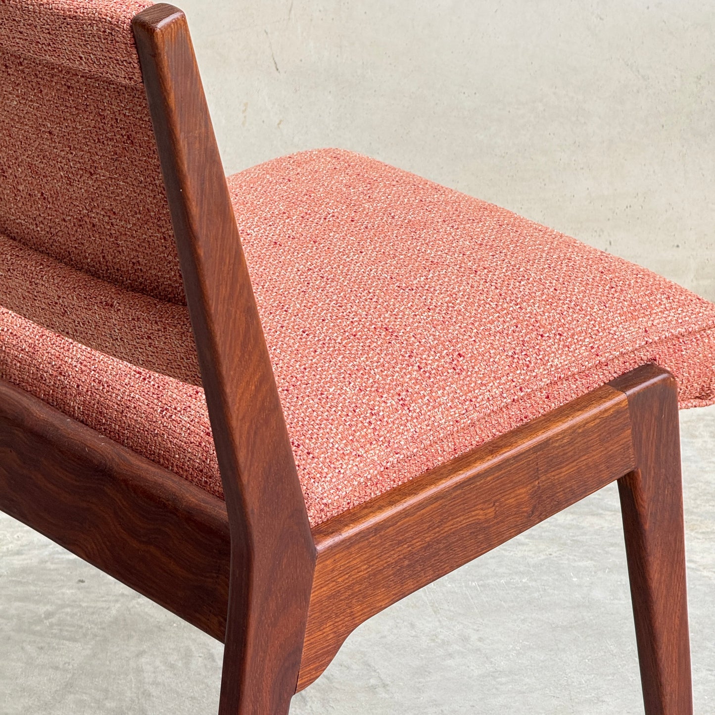 A close-up of a Vintage MCM Jens Risom Chair from a collectable set of 4 by Refine, showing its reddish-pink textured upholstery and smooth, angled wooden frame against a plain, light-colored background.