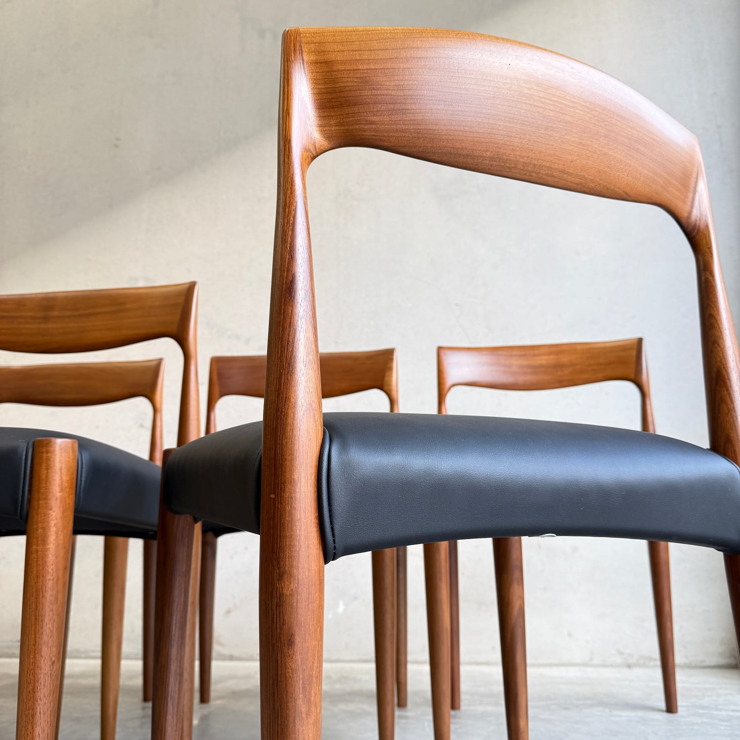 Four modern wooden chairs with curved backrests and black upholstered seats are arranged in a minimalist setting against a plain, light-colored wall.