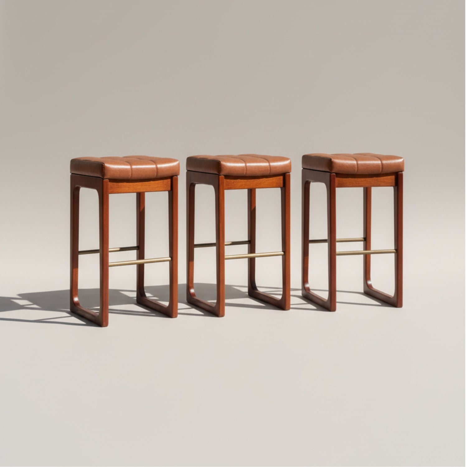 Three modern wooden bar stools with square padded brown leather seats and brass footrests are arranged in a row on a neutral background.