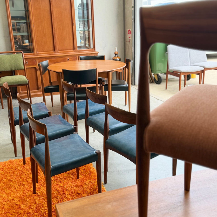 A collection of mid-century modern wooden chairs and tables, some with blue or brown upholstery, arranged indoors on a concrete floor with an orange shag rug.