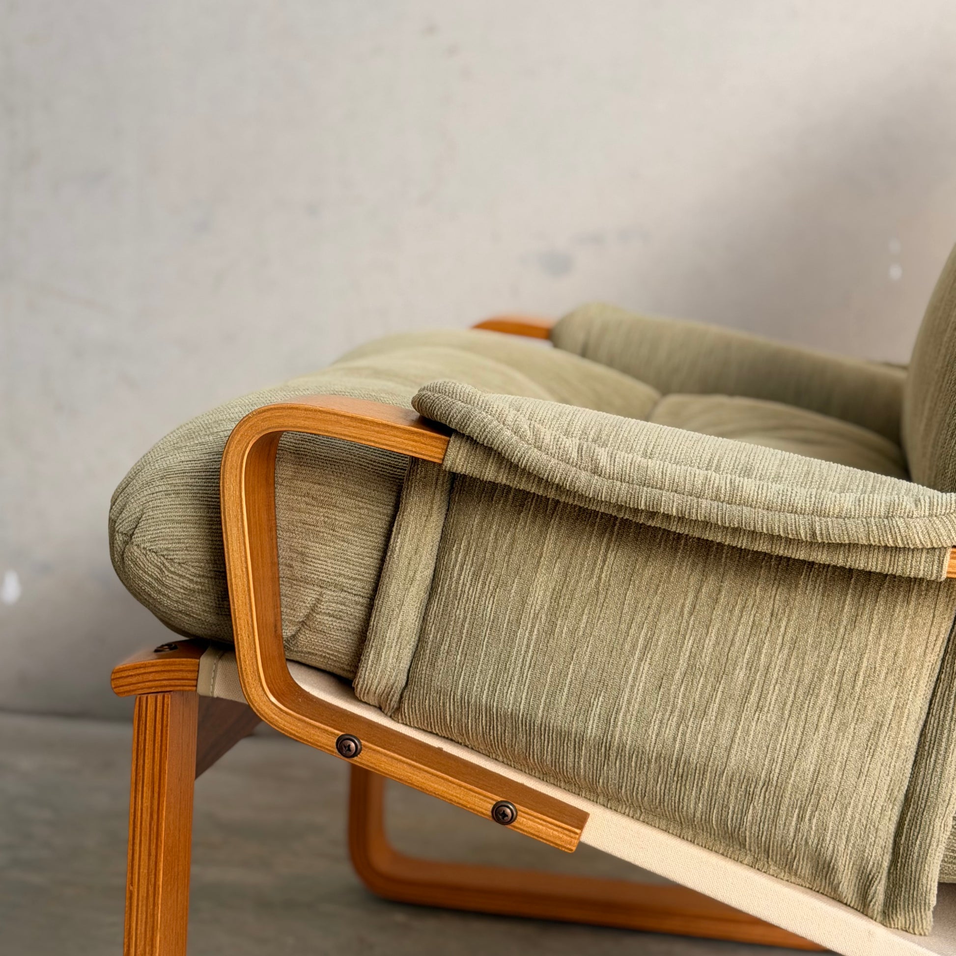 A close-up of a Pair of Tessa Monaco Arm Chairs - Sage Green Corduroy Like New (Revive), featuring textured sage cushions and curved wooden armrests, set against a plain light gray wall and concrete floor.