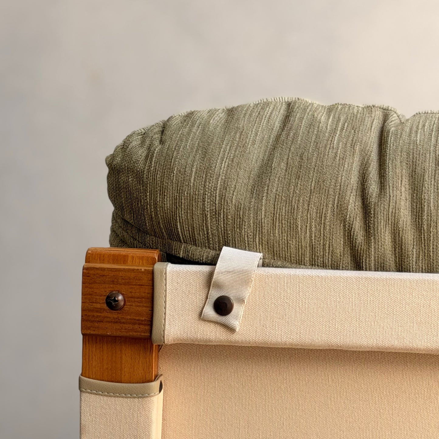 Close-up of a Tessa Monaco Arm Chair—sage green corduroy cushion and beige canvas backrest, strap secured by a dark button. The plain background and elegant design evoke mid-century style. Pair of chairs, like new (Revive).
