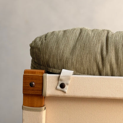 Close-up of a Tessa Monaco Arm Chair—sage green corduroy cushion and beige canvas backrest, strap secured by a dark button. The plain background and elegant design evoke mid-century style. Pair of chairs, like new (Revive).