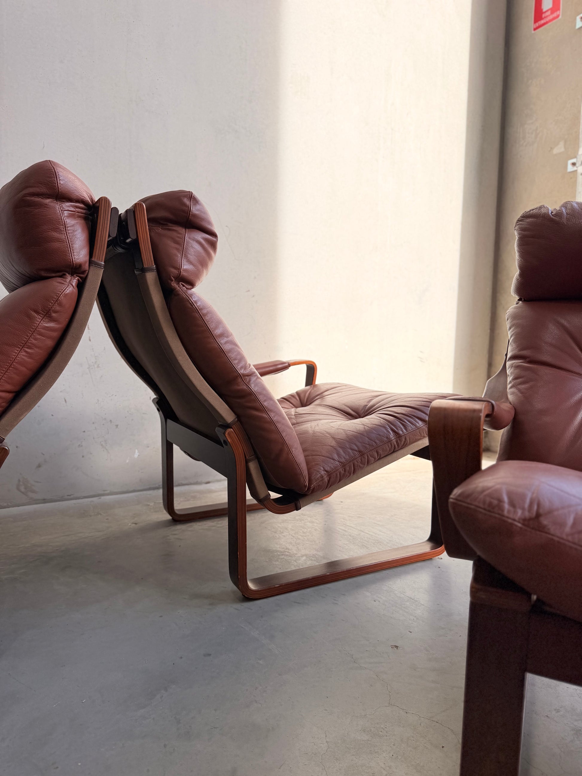 A Tessa T8 Lounge Suite—Revive modular two-seater sofa and armchairs in brown leather, designed by Fred Lowen in mid-century modern style, are arranged on a concrete floor with sunlight casting soft shadows near a beige wall.