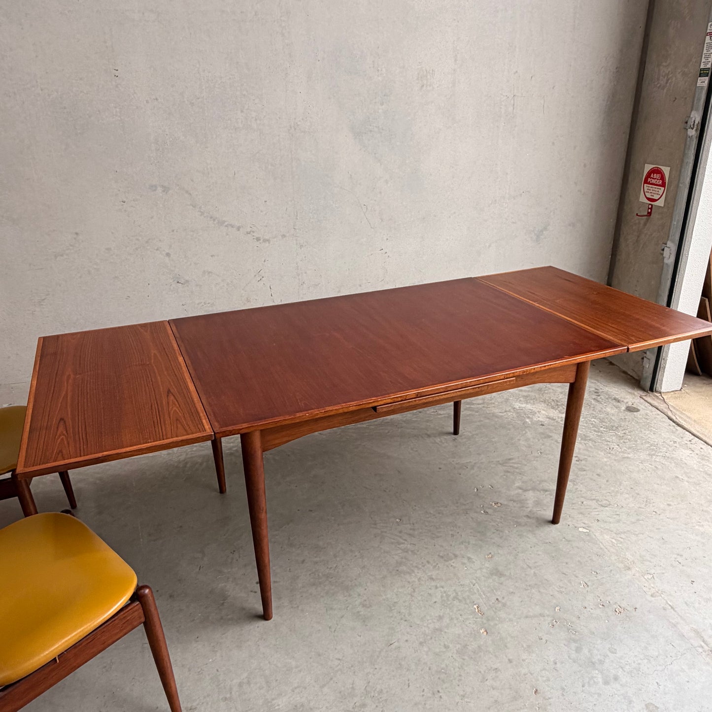 A Vintage Mid-Century Parker 109 Nordic Extension Dining Table (table only) with tapered legs sits on a concrete floor near a plain light wall, with part of a yellow-cushioned chair visible on the left.