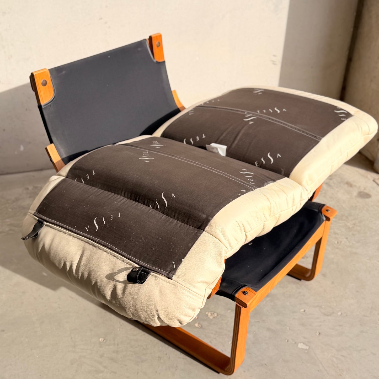 The Tessa Austin Blonde Leather Armchair (Revive) features a wooden frame and thick, patterned black-and-beige cushion, embodying mid-century modern style and elegance, perfect for sunlit spaces with concrete floors.