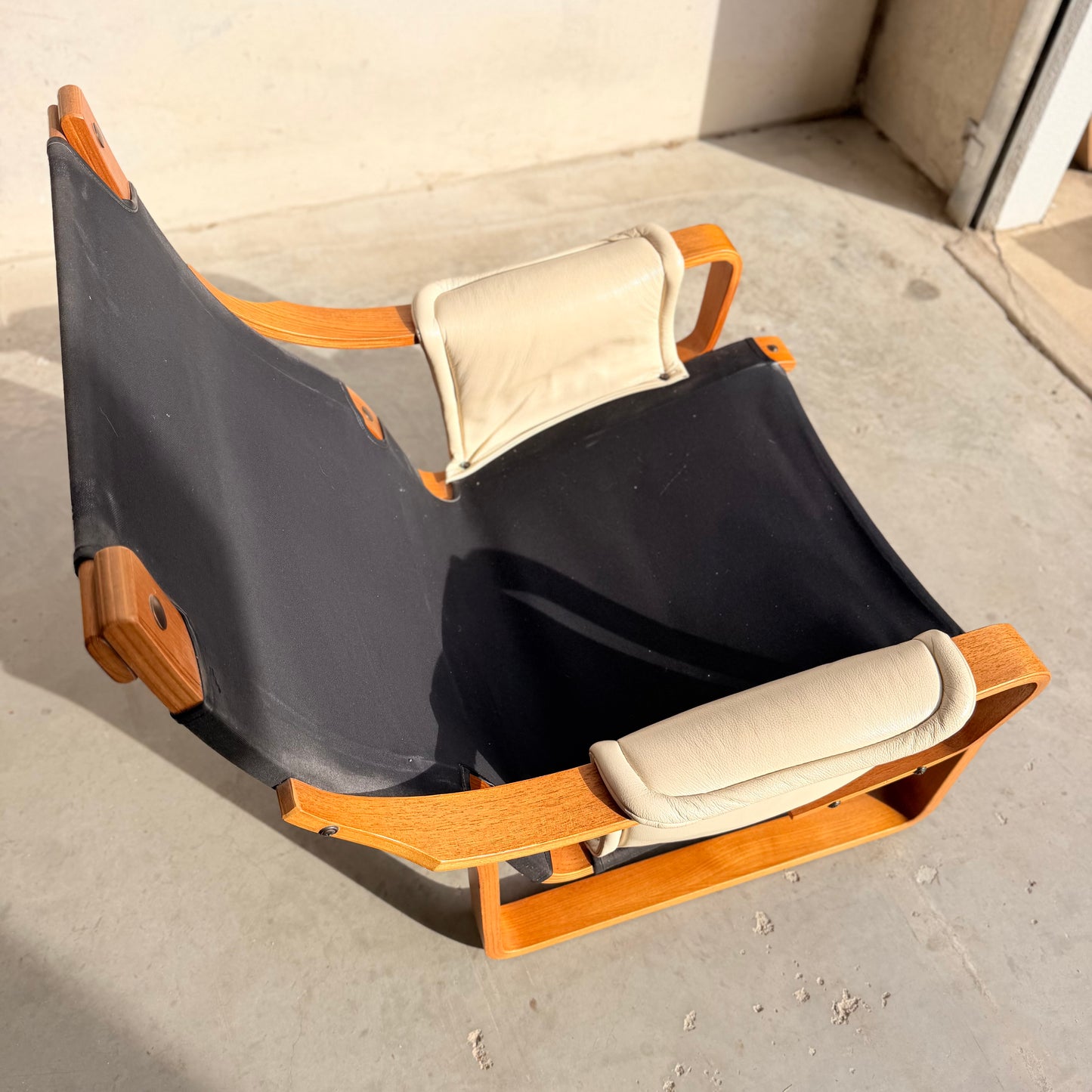 The Tessa Austin Blonde Leather Armchair (Revive) features a low wooden frame, black canvas seat, and two light beige cushions, evoking mid-century modern style in bright sunlight on a concrete floor near a wall.