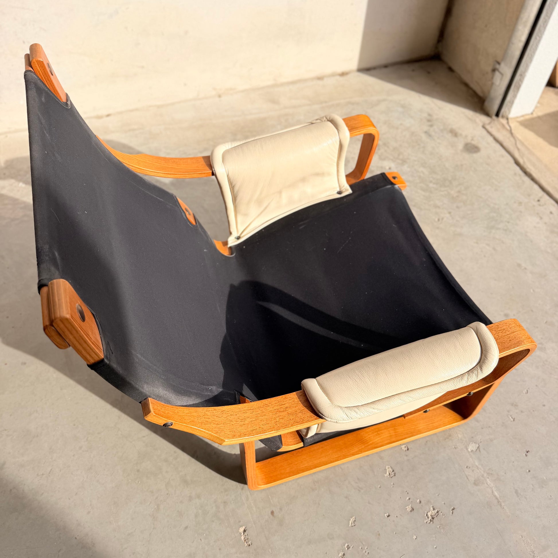 The Tessa Austin Blonde Leather Armchair (Revive) features a low wooden frame, black canvas seat, and two light beige cushions, evoking mid-century modern style in bright sunlight on a concrete floor near a wall.