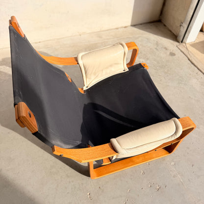 The Tessa Austin Blonde Leather Armchair (Revive) features a low wooden frame, black canvas seat, and two light beige cushions, evoking mid-century modern style in bright sunlight on a concrete floor near a wall.