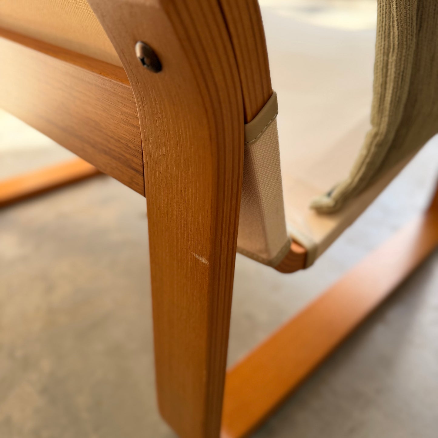 Close-up of a Tessa Monaco Arm Chair in sage green corduroy, showing its curved wooden leg, beige fabric strap, and textured seat. Inspired by mid-century design. Pair of chairs available like new from Revive. Background is out of focus.