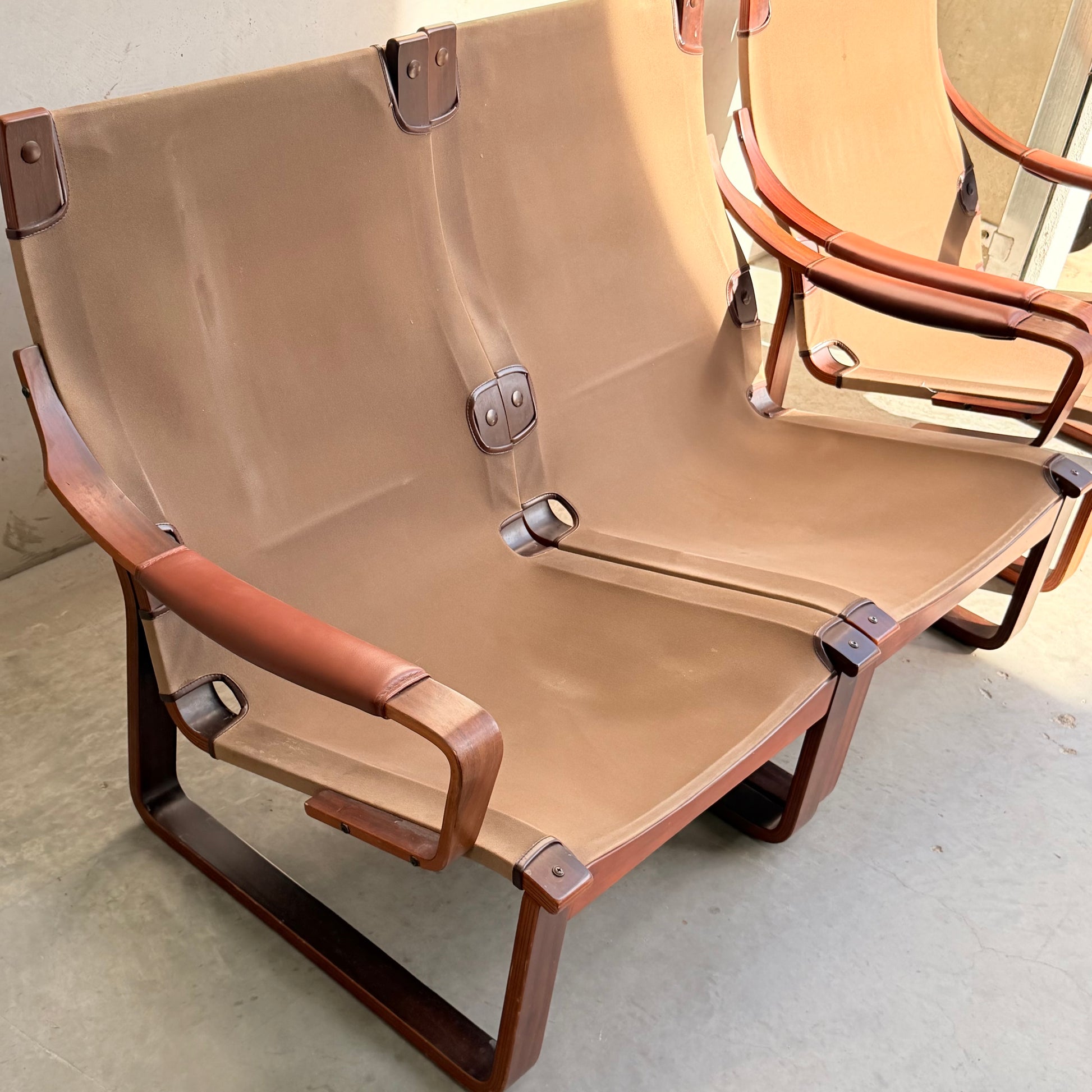 A modular Tessa T8 Lounge Suite, featuring a two-seater sofa and armchairs in modern brown canvas with wood frames and leather accents, sits on a concrete floor beside a light-colored wall.