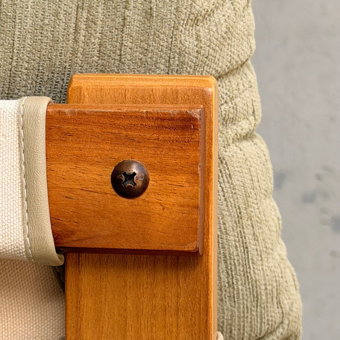 Close-up of a wooden chair with a metal screw, beige cushion, and light fabric strap set against gray—ideal to pair with the Pair of Tessa Monaco Arm Chairs - Sage Green Corduroy Like New (Revive).