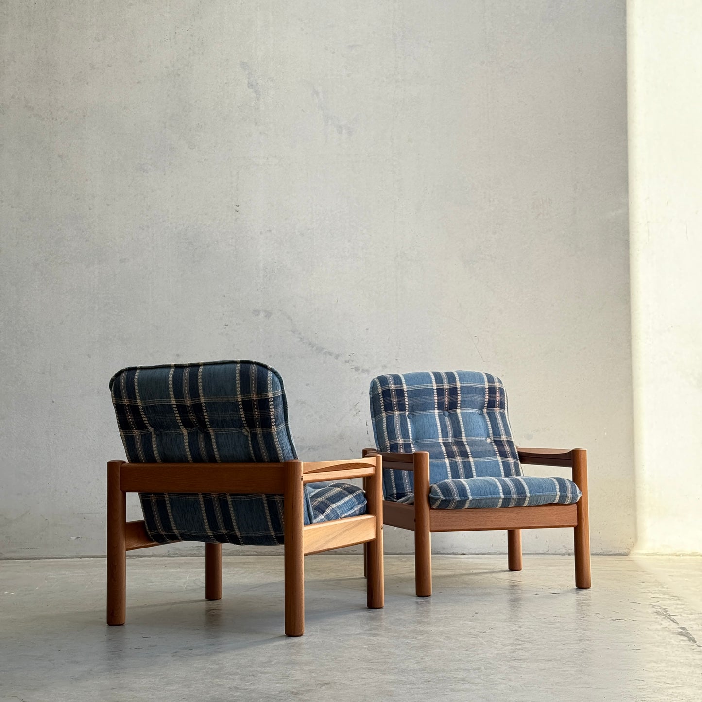 Two Restored Domino Møbler Armchairs from the Blue Plaid Mid-Century Set (Rediscover) sit on a concrete floor by a light wall, bathed in natural light from the right.