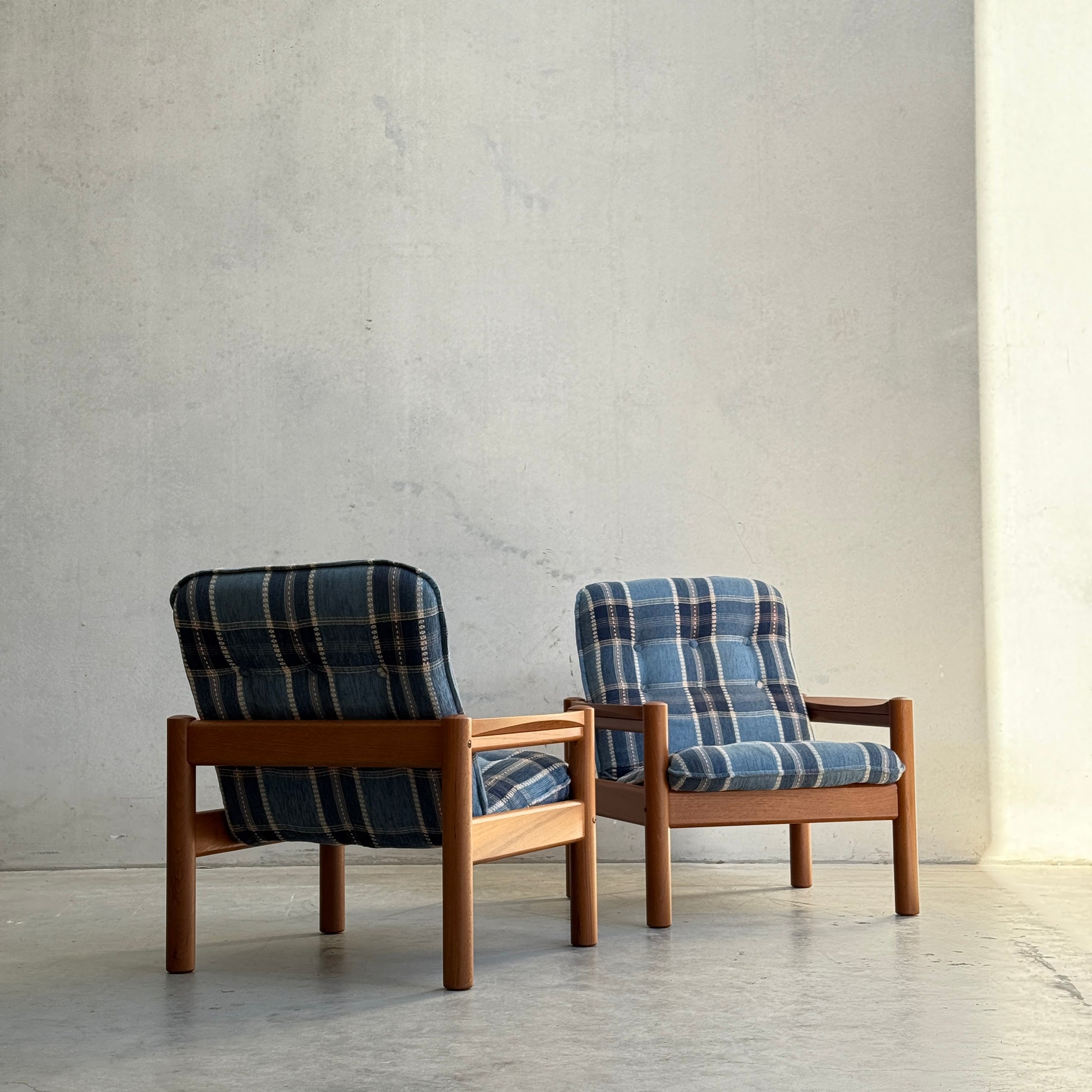 Two Restored Domino Møbler Armchairs from the Blue Plaid Mid-Century Set (Rediscover) sit on a concrete floor by a light wall, bathed in natural light from the right.