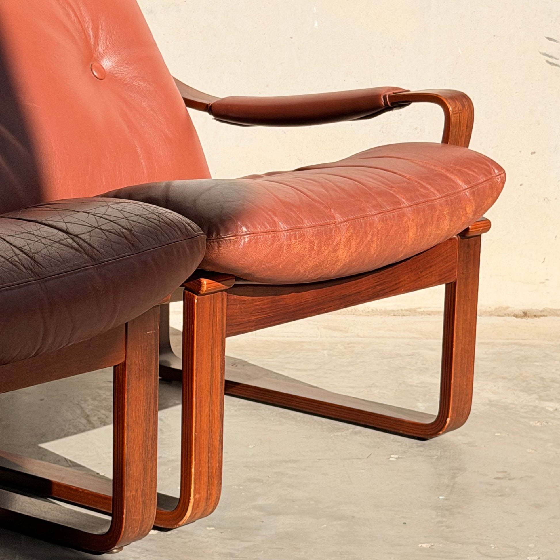 Two Tessa T8 Lounge Suite (Revive) modular armchairs, with reddish-brown leather cushions and wooden frames, sit side by side on a sunlit concrete floor.