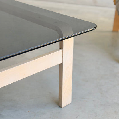 A close-up of the Tessa Ferrara Lounge Suite – Complete 5-Piece Set (Revive), featuring a black glass top and light wood legs, rests on a smooth light-gray floor, capturing the timeless elegance of mid-century modern design.