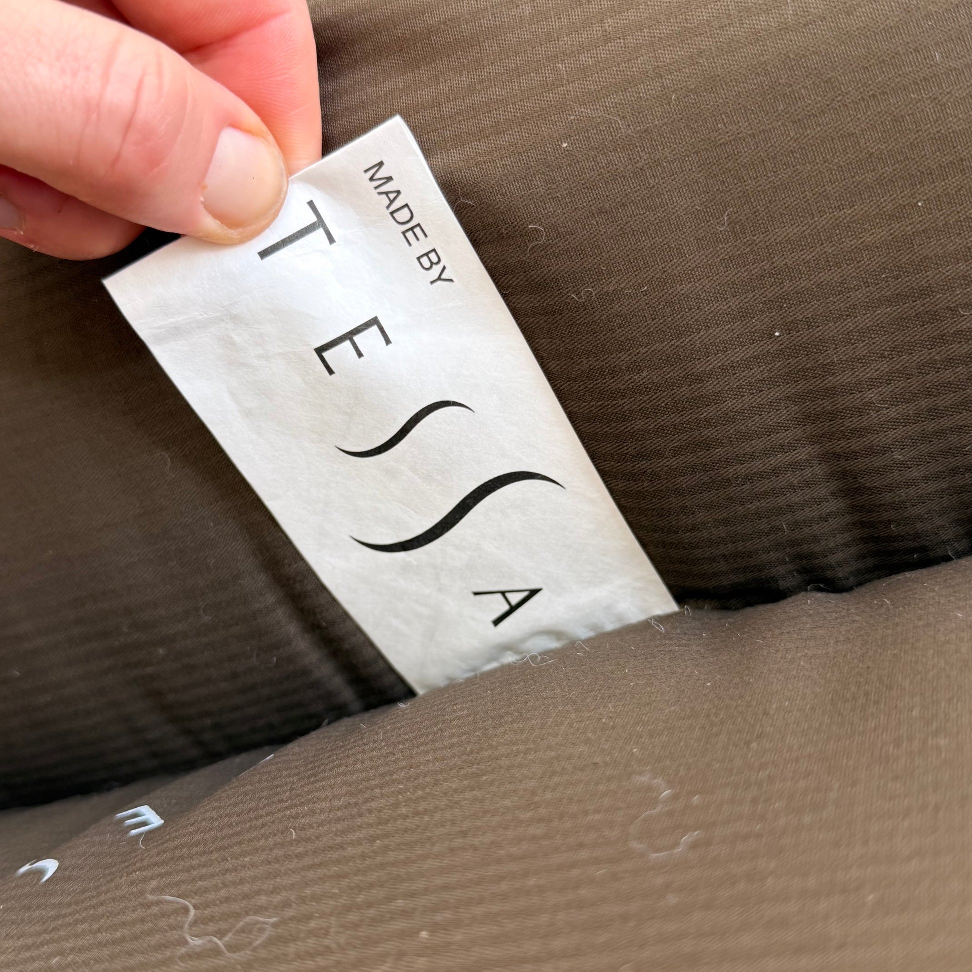A hand holds a white MADE BY TESSA tag on a brown, ribbed cushion, with visible lint, from a Pair of Tessa Monaco Arm Chairs - Sage Green Corduroy Like New (Revive), inspired by mid-century design.