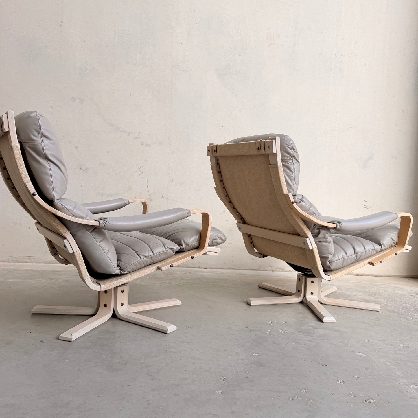 The Tessa Ferrara Lounge Suite – Complete 5-Piece Set (Revive) features light wood frames and gray upholstery; two chairs are set on a concrete floor against a plain wall, one facing forward and the other displaying its back.
