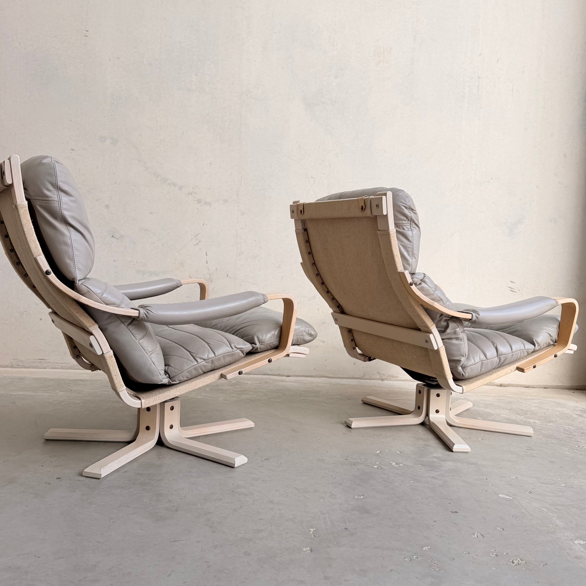 The Tessa Ferrara Lounge Suite – Complete 5-Piece Set (Revive) features light wood frames and gray upholstery; two chairs are set on a concrete floor against a plain wall, one facing forward and the other displaying its back.