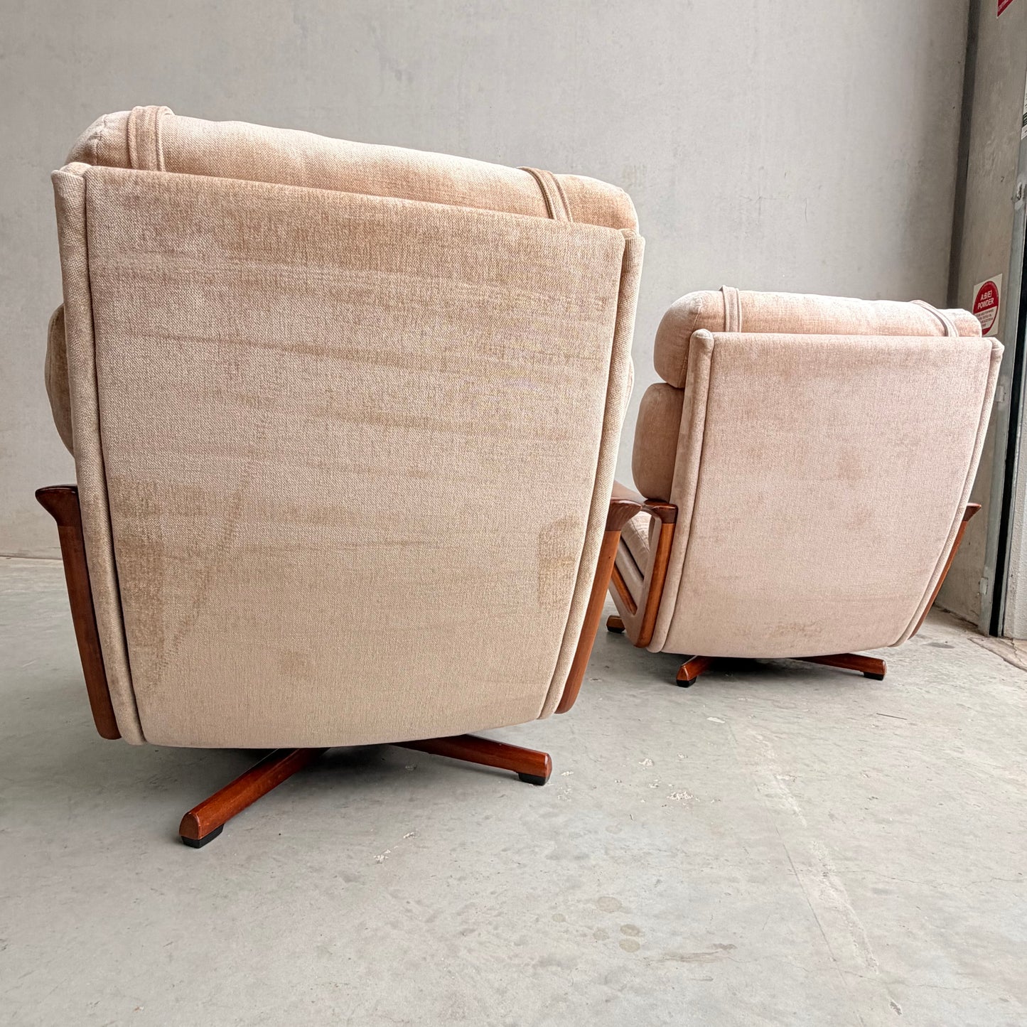 Two light beige Vintage Mid Century Tessa T21 Swivel Chairs (Revive) with wooden bases sit on a concrete floor, facing away from the camera toward a blank, light-colored wall.