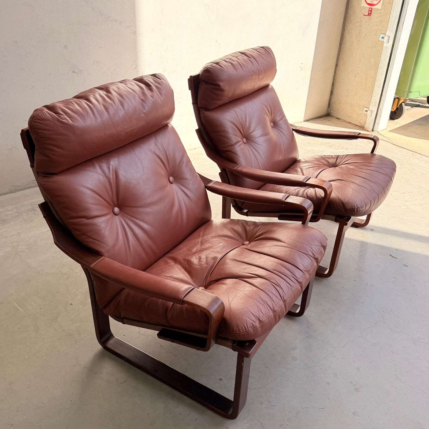 The Tessa T8 Lounge Suite modular two-seater sofa and armchairs (Revive), featuring brown leather and curved wooden frames, sit side by side in a sunlit room with a concrete floor, evoking mid-century modern style.