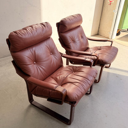 The Tessa T8 Lounge Suite modular two-seater sofa and armchairs (Revive), featuring brown leather and curved wooden frames, sit side by side in a sunlit room with a concrete floor, evoking mid-century modern style.