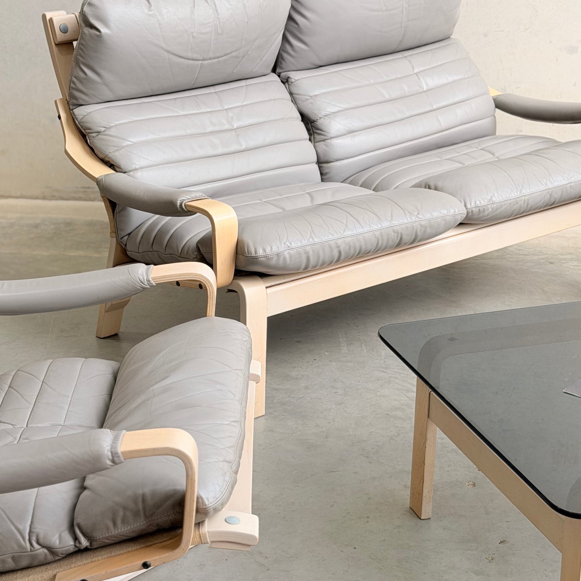 The Tessa Ferrara Lounge Suite – Complete 5-Piece Set (Revive) by Fred Lowen features light wood frames and padded gray leather cushions, arranged on a polished concrete floor beside a glass coffee table with a wooden base.
