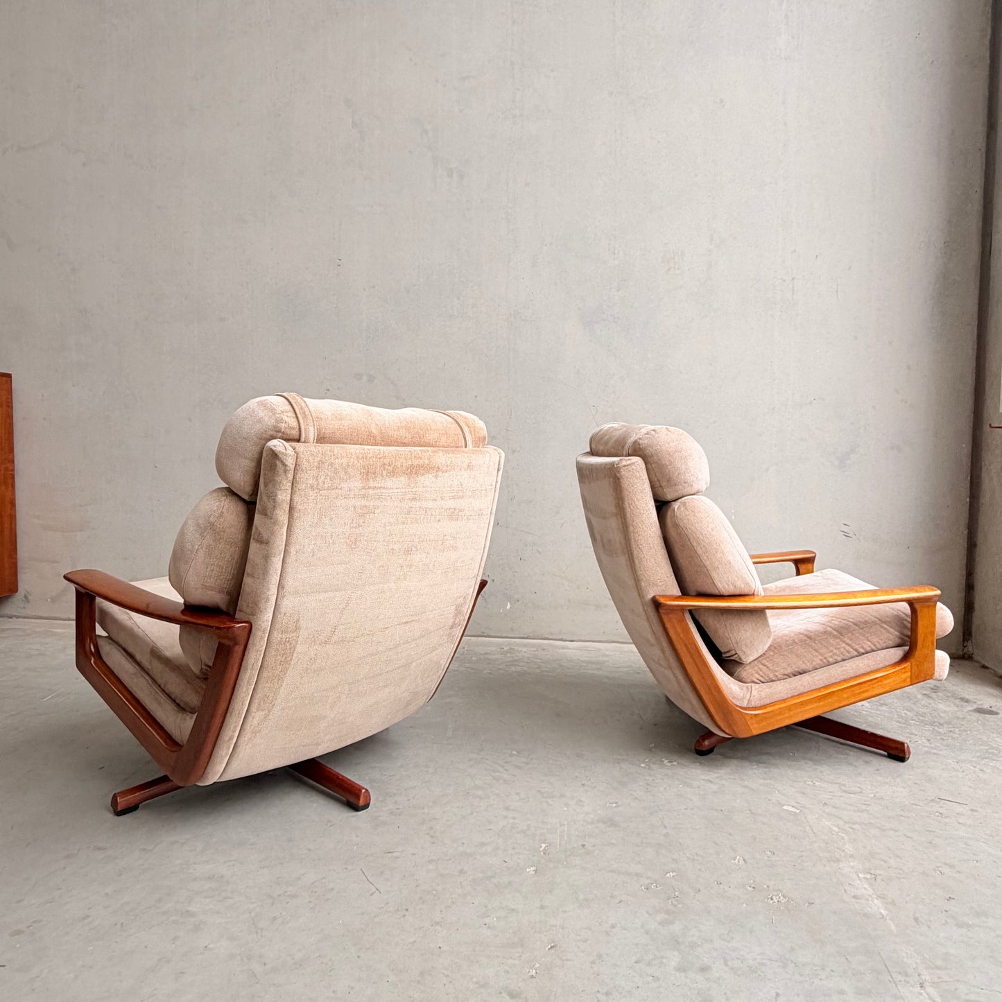 Two Vintage Mid Century Tessa T21 Swivel Chairs (Revive) with cream velvet upholstery and wooden frames sit on a concrete floor against a plain, light wall, positioned to highlight their elegant mid-century design and swivel feature.