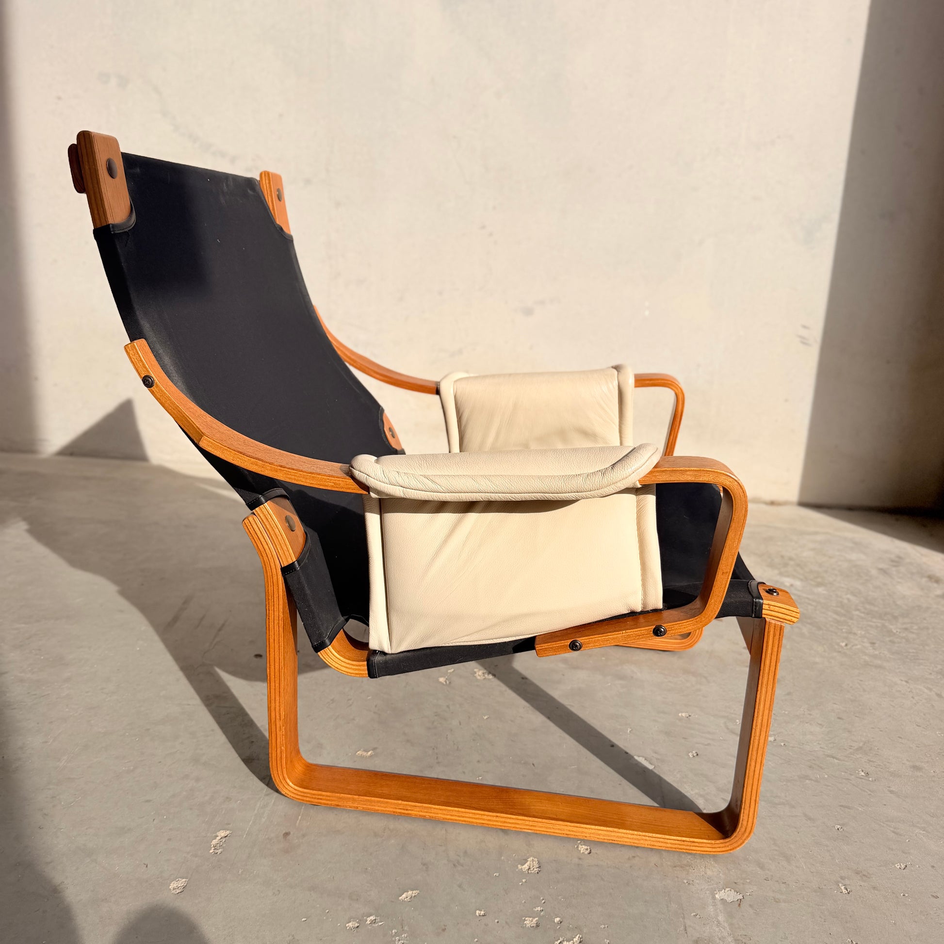 The Tessa Austin Blonde Leather Armchair (Revive) features a curved wooden frame, black fabric seat, and light beige cushions, capturing mid-century modern elegance on a concrete floor against a plain light wall.