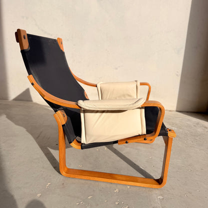 The Tessa Austin Blonde Leather Armchair (Revive) features a curved wooden frame, black fabric seat, and light beige cushions, capturing mid-century modern elegance on a concrete floor against a plain light wall.