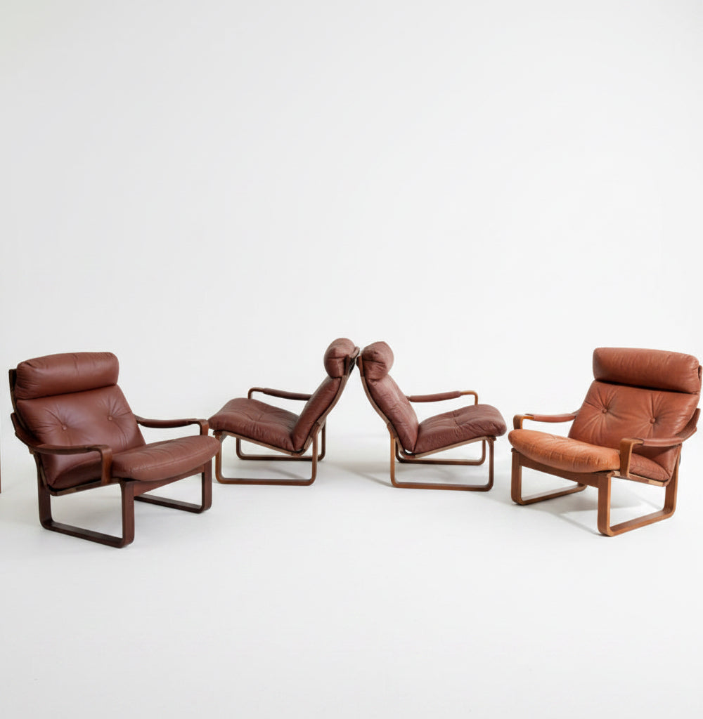 Four Tessa T8 Lounge Suite modular two-seater sofas and armchairs (Revive) in brown leather with wooden frames, inspired by mid-century modern design, are arranged in a minimalist, sunlit room with light grey walls and a pale floor. A wooden cabinet sits in back.