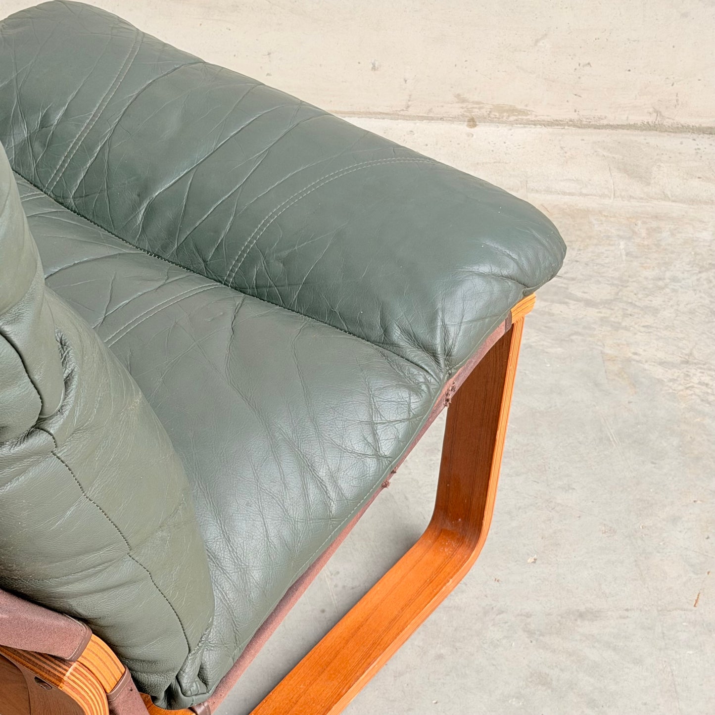 Close-up of the Tessa Vintage Single No Arm Monaco Armchair – Forest Green (Revive) with leather cushion and wooden frame, set on a light concrete floor against a plain wall.