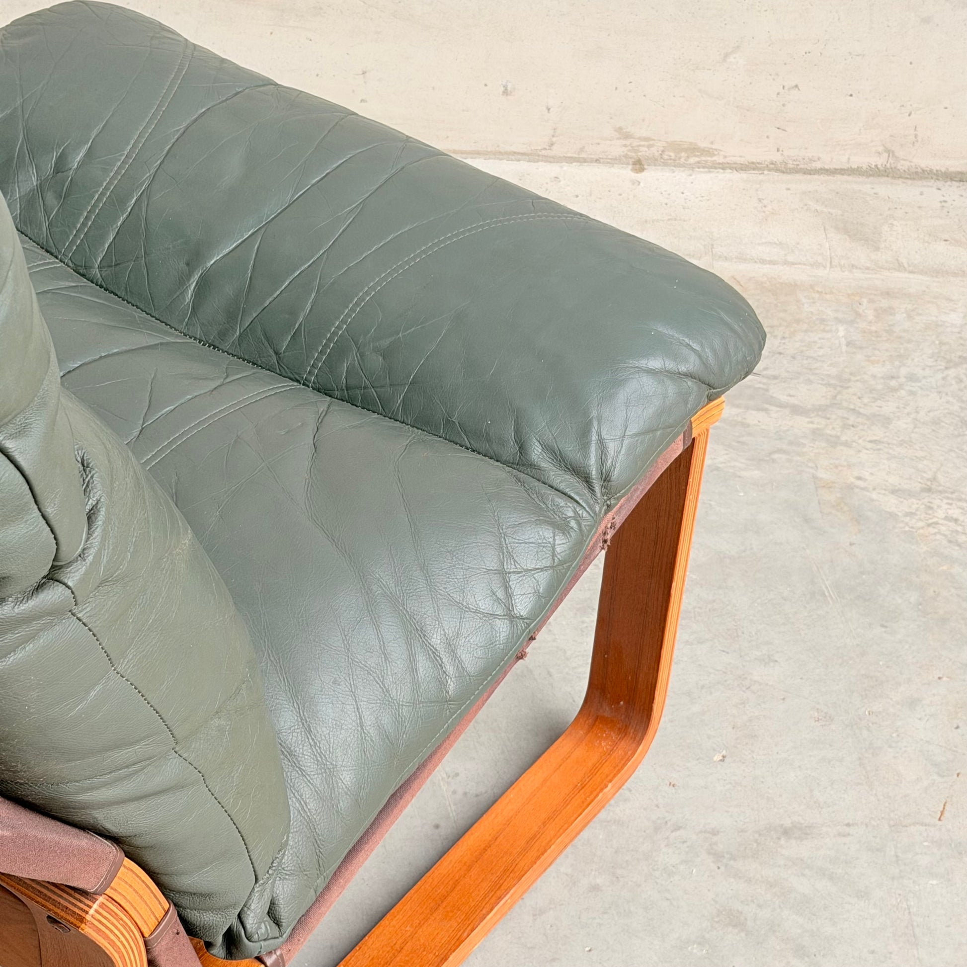 Close-up of the Tessa Vintage Single No Arm Monaco Armchair – Forest Green (Revive) with leather cushion and wooden frame, set on a light concrete floor against a plain wall.