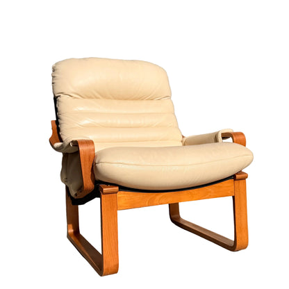 The Tessa Austin Blonde Leather Armchair (Revive), featuring a wooden frame and armrests, sits on a concrete floor against a plain light wall, illuminated by natural sunlight.