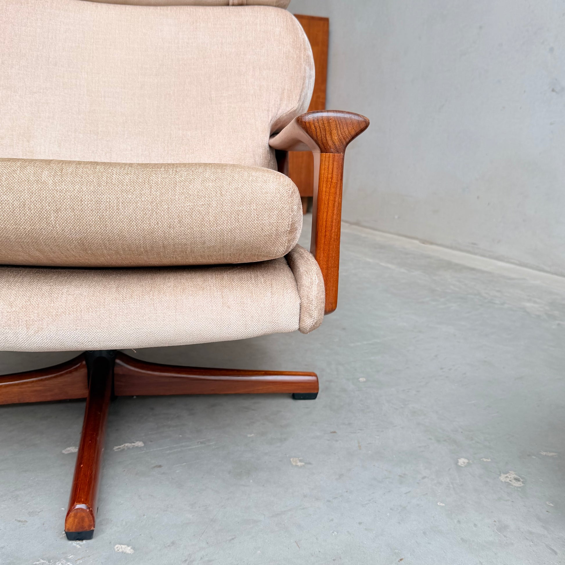 Close-up of the beige upholstered Vintage Mid Century Tessa T21 Swivel Chair (Revive) with wooden armrests and legs, set on a grey concrete floor against a light wall. The partial view highlights its iconic mid-century design.