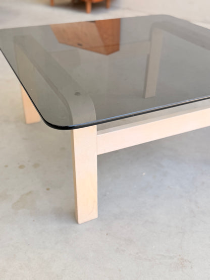 A square coffee table with light wood legs and a dark tinted glass top, inspired by mid-century style, pairs beautifully with the Tessa Ferrara Lounge Suite – Complete 5-Piece Set (Revive) on a light-colored floor.