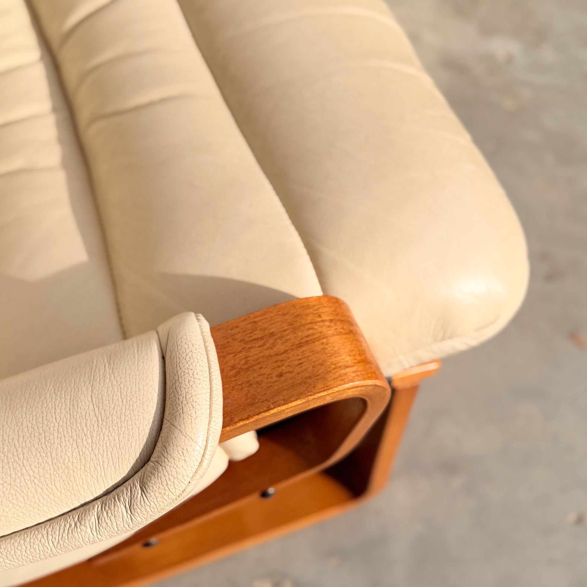 Close-up of the Tessa Austin Blonde Leather Armchair (Revive), featuring a cream leather cushion with visible stitching on a wooden frame; sunlight highlights its mid-century modern textures.