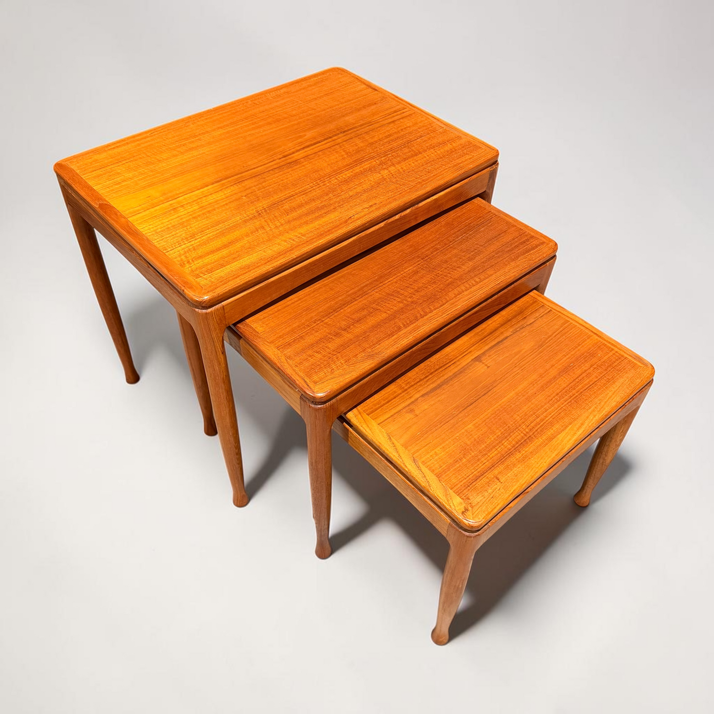 A rare set of three Alpa Teak Nesting Tables, Australian Mid-Century Modern by Revive, feature tapered legs and a warm brown finish, each table partially fitting under the next against a plain light background.