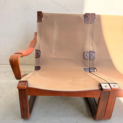 A close-up of the Tessa T8 Lounge Suite modular two-seater sofa and armchairs (Revive), showcasing a wooden frame, tan canvas seat and backrest, leather straps, and metal fastenings, set against a light-colored wall and floor.